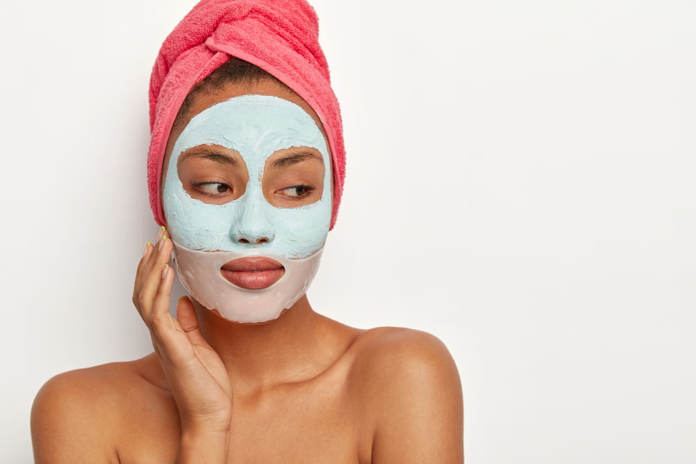 people beauty self care concept thoughtful dark skinned woman touches cheek applies clay mask collagen patch around lips wears towel head